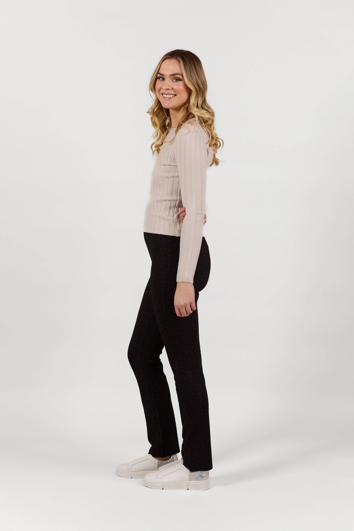 A young woman with long wavy blonde hair stands smiling sideways, wearing a light ribbed sweater, Vassalli Bay Pull On - Sprig slim leg pants, and white sneakers against a plain white background.