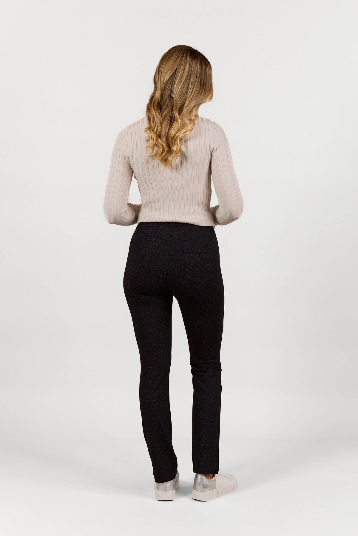 A woman with long, wavy blonde hair stands facing away against a white background, wearing Vassalli's Bay Pull On - Sprig slim leg pants in stretch fabric paired with a light beige ribbed sweater and light-colored shoes.