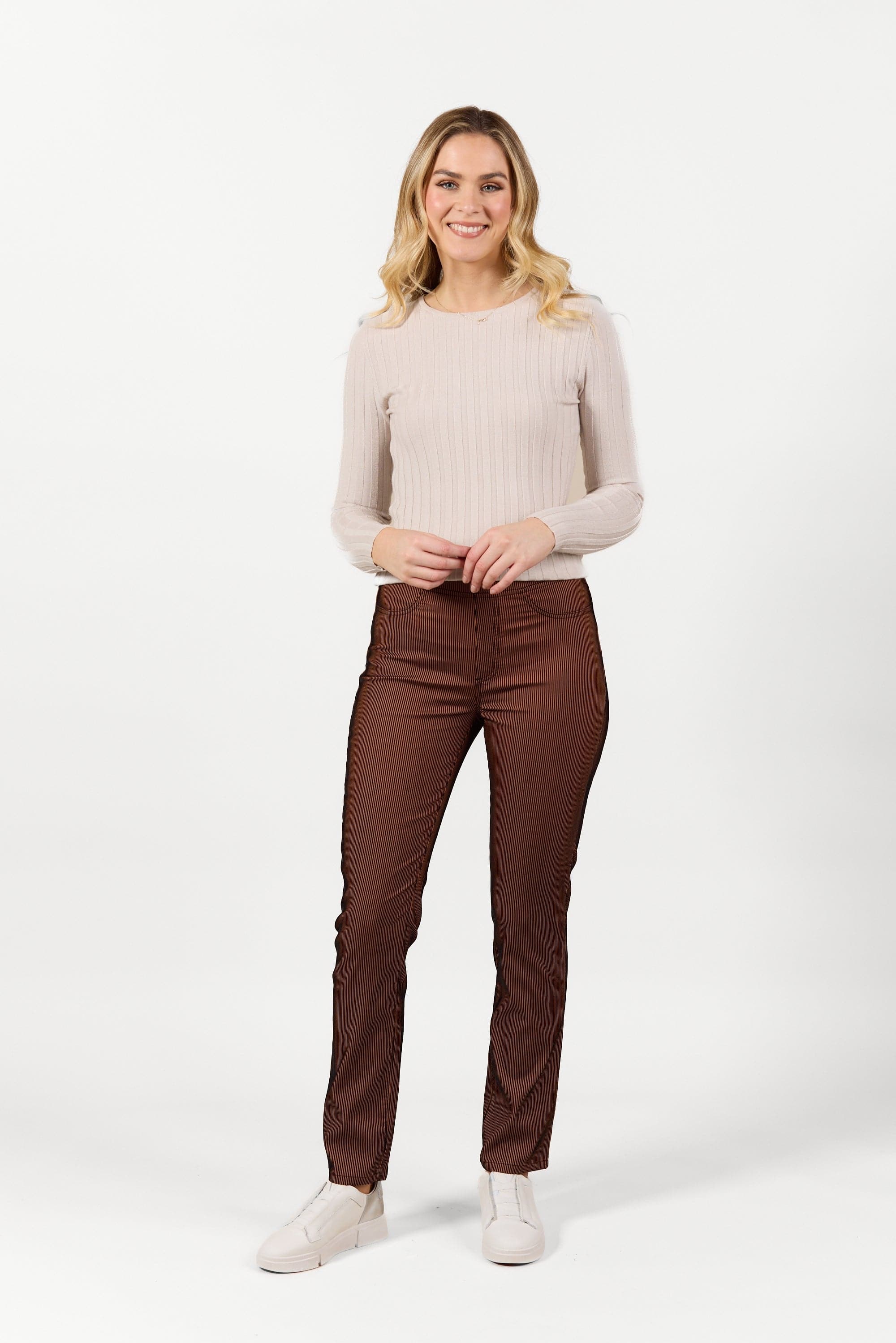 A woman with long blonde hair, in a light beige ribbed sweater and Vassalli's Bay Pull On - Tiger Velvet pants, smiles while standing against a plain white background.