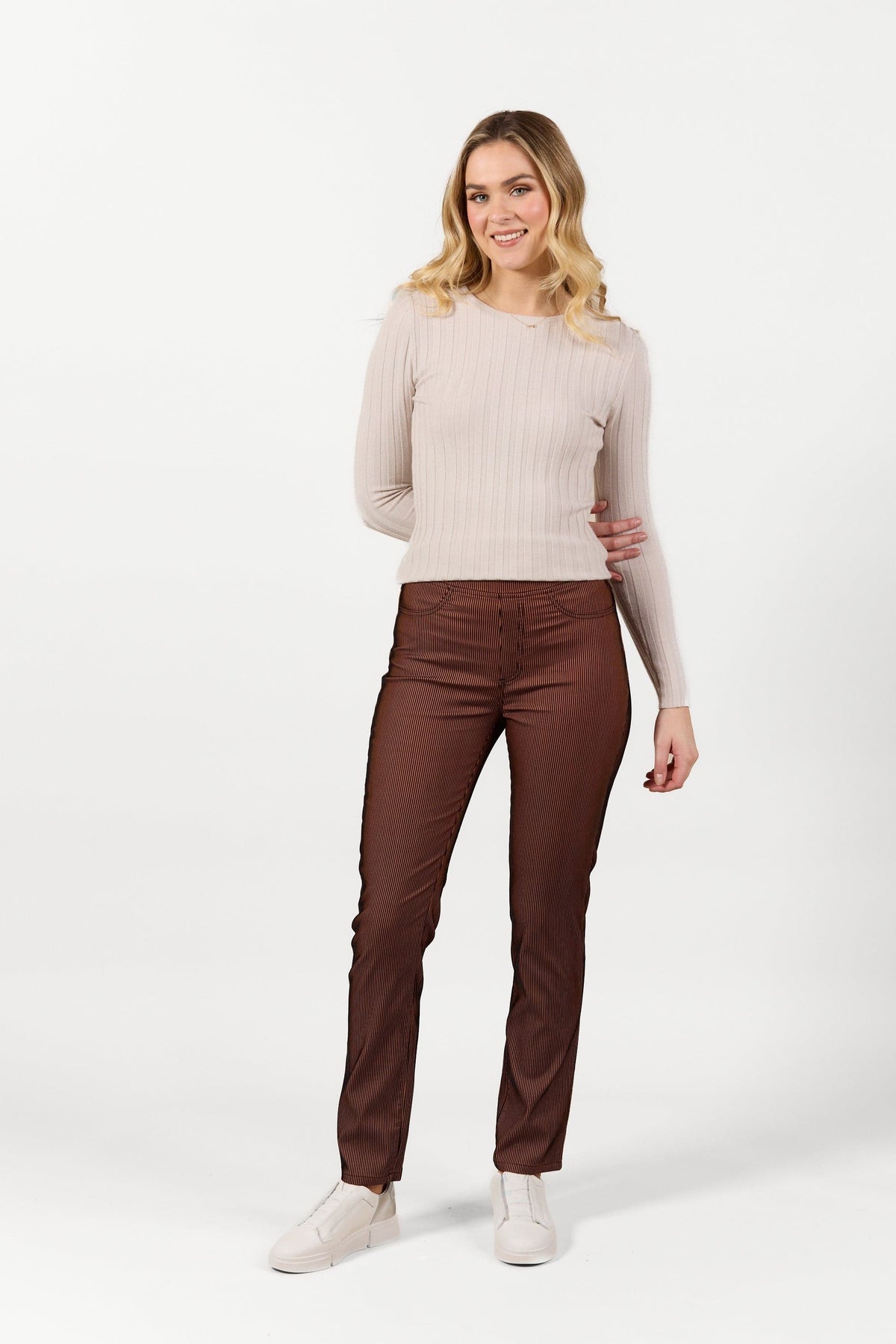 A woman with blonde hair stands smiling, wearing a Vassalli Bay Pull On - Tiger Velvet pants and a light beige long-sleeve ribbed top with white sneakers, against a plain white background.