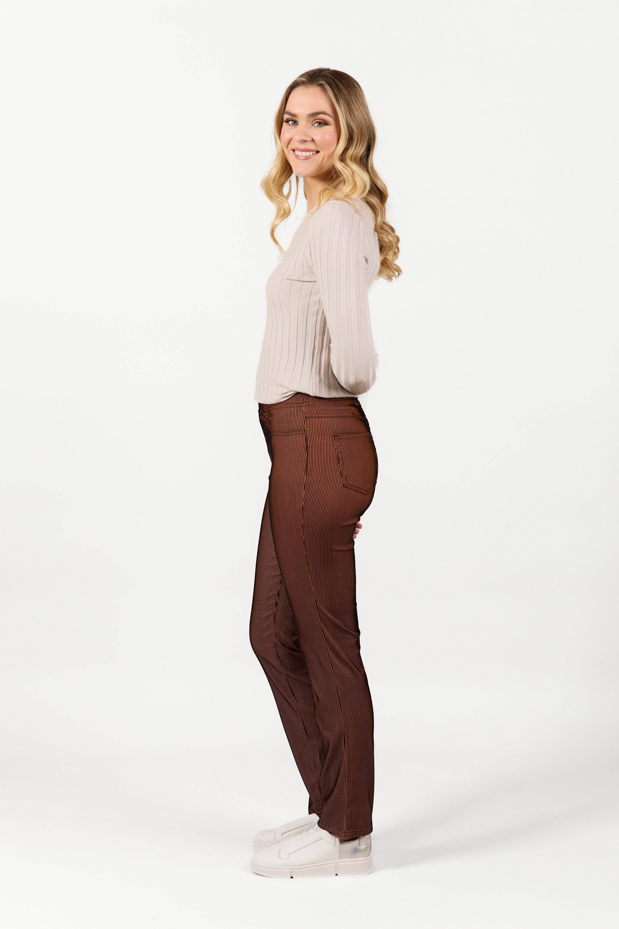A young woman with long, wavy blonde hair stands sideways, smiling. She wears a cream long-sleeve top and Vassalli Bay Pull On - Tiger Velvet pants with an elastic waistband, paired with white platform shoes against a plain white background.