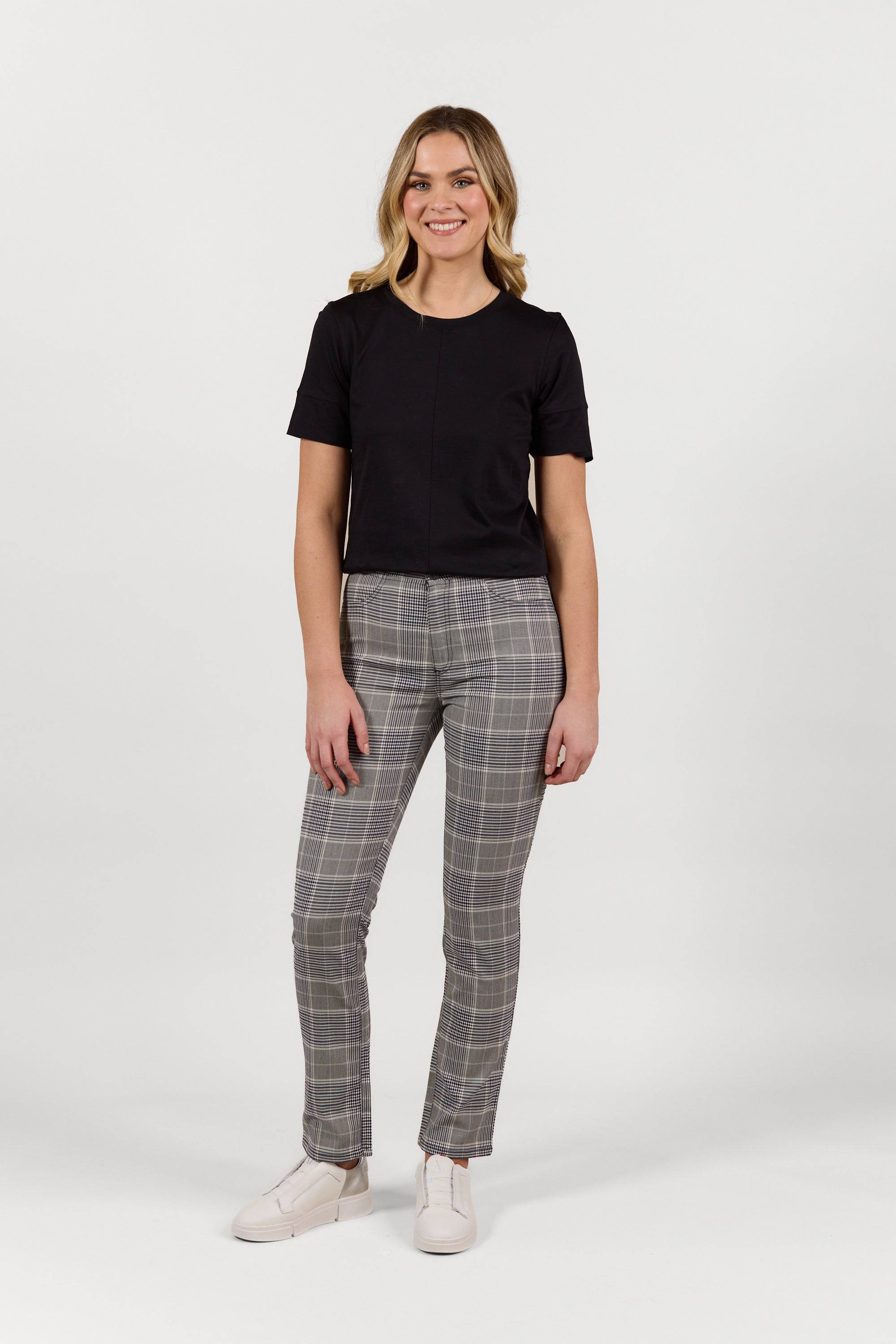 A woman smiles, wearing a black short-sleeve top, Vassalli Bay Pant - Danbury in gray plaid with an elastic waistband, and white sneakers against a plain white background.
