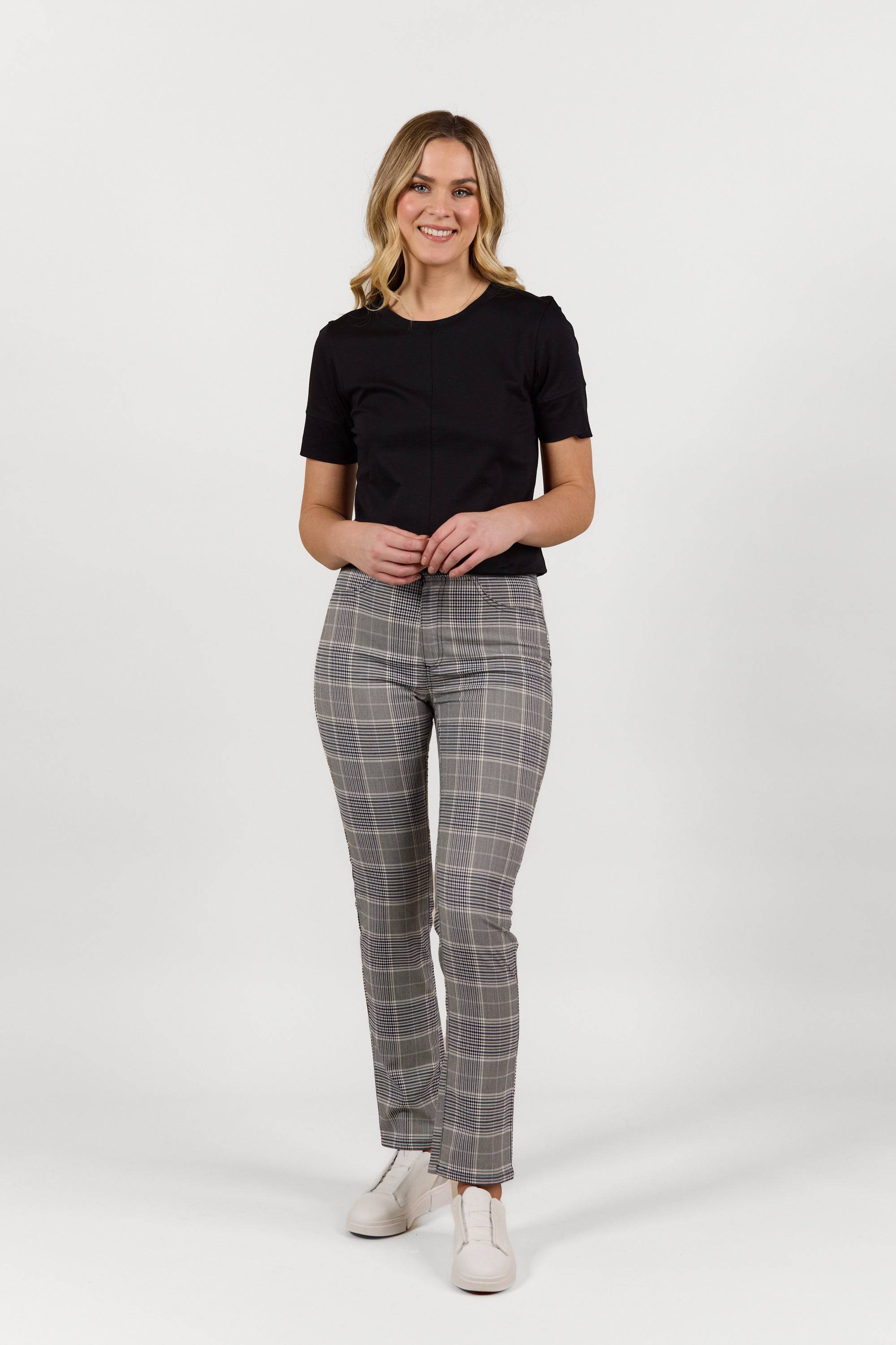 A woman with light wavy hair smiles against a plain white background, wearing the Vassalli Bay Pant - Danbury, a gray plaid slim leg pant with an elastic waistband, paired with a black short-sleeve top and white shoes. Her hands are lightly clasped.