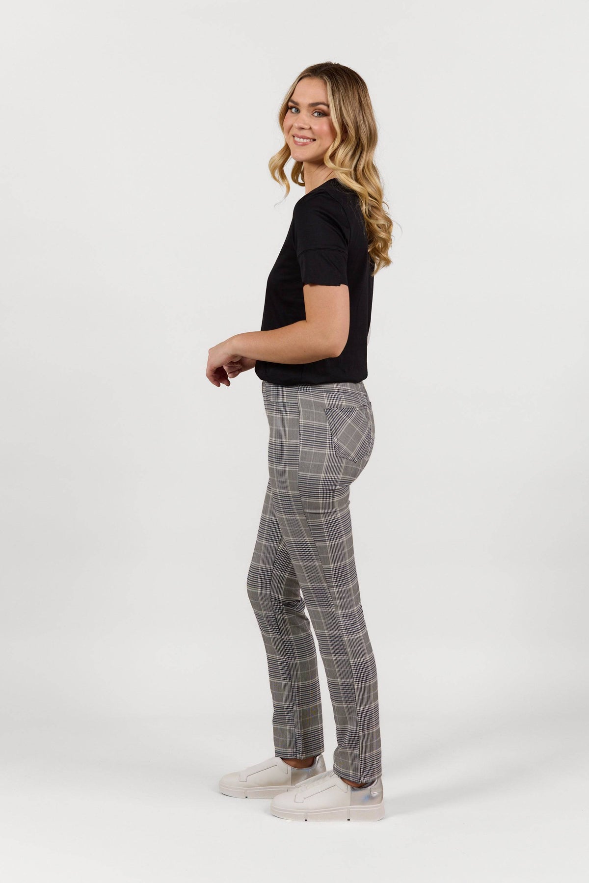 A woman with long, wavy blonde hair stands sideways, smiling. She wears Vassalli’s Bay Pant - Danbury with an elastic waistband, a black t-shirt, and white sneakers against a plain white background.