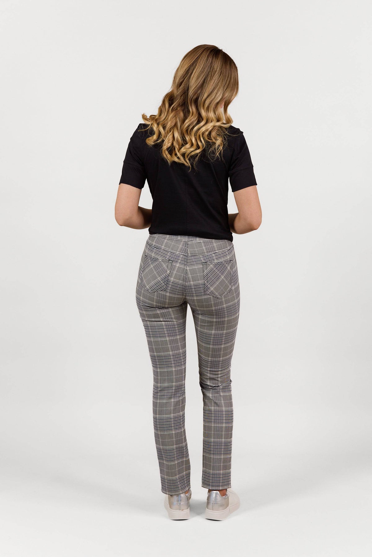 A woman with wavy blonde hair wears a black short-sleeve top and Vassalli Bay Pant - Danbury gray plaid slim leg pants with an elastic waistband, standing facing away from the camera against a plain white background.