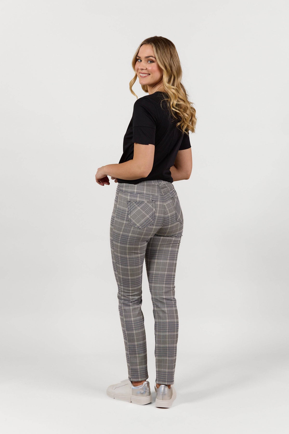 A woman with long wavy hair smiles over her shoulder, wearing a black t-shirt and white sneakers paired with Vassalli's Bay Pant - Danbury gray plaid slim leg pants with an elastic waistband, set against a plain white background.