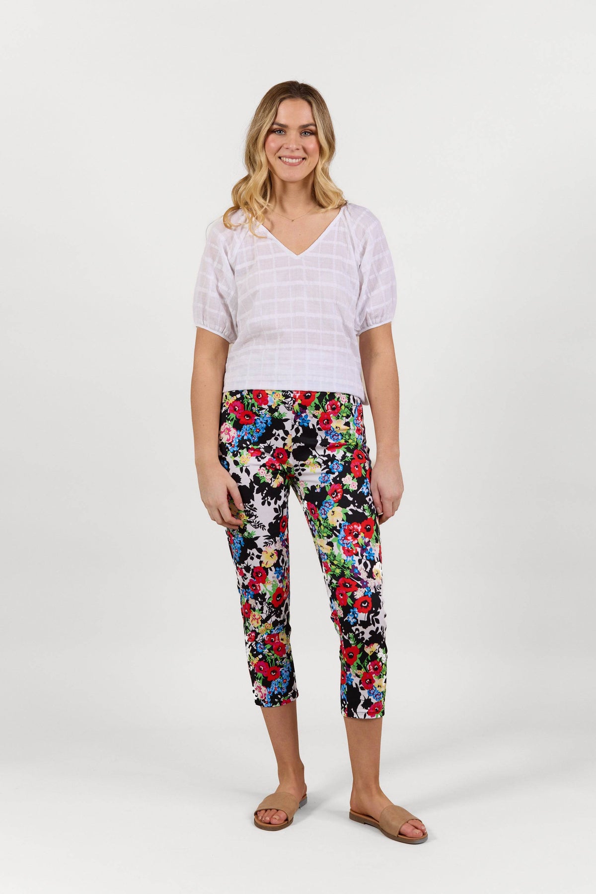 A woman smiles, wearing a white short-sleeve V-neck blouse, Vassalli Harbour 7/8 Pant - Bouquet, and tan sandals against a plain white background.