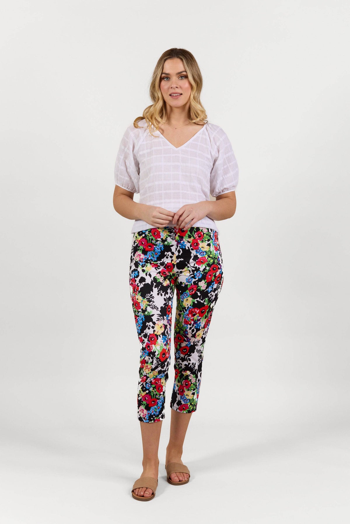 A woman stands against a white background wearing a white short-sleeve blouse, Vassalli Harbour 7/8 Pant - Bouquet, and tan sandals. She faces forward with her hands loosely clasped.