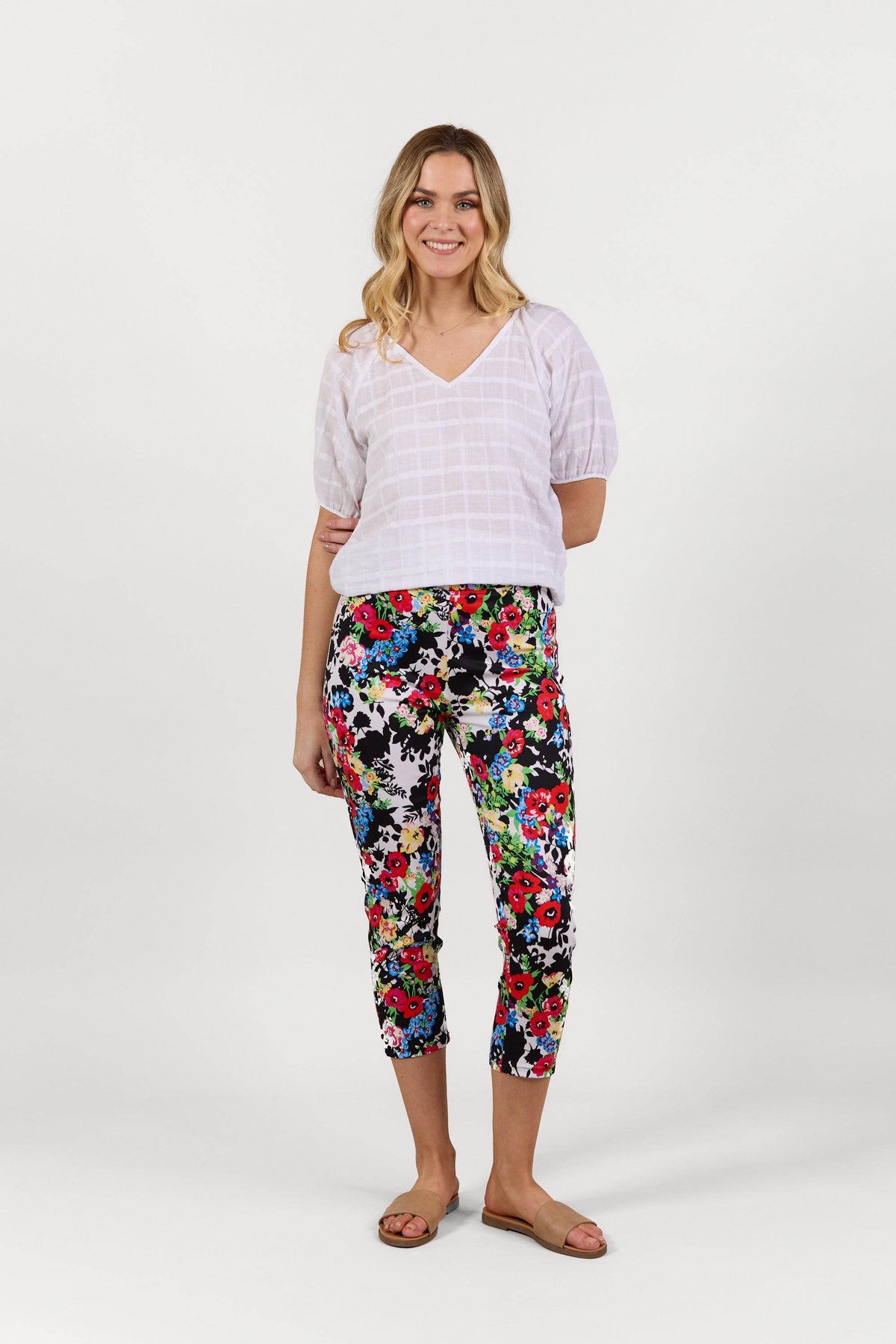 A smiling woman stands against a white background, wearing a white short-sleeve blouse, Vassalli Harbour 7/8 Pant - Bouquet in stretch fabric, and tan sandals.