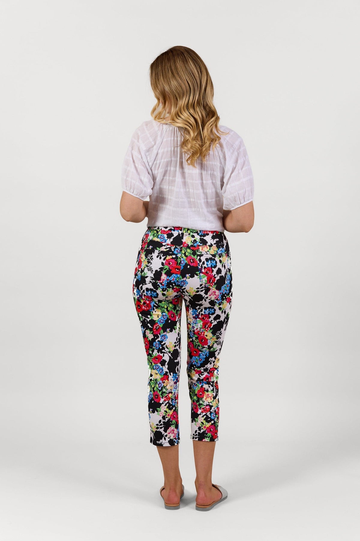 A woman with wavy blonde hair faces away, wearing a white blouse and Vassalli Harbour 7/8 Pant - Bouquet, featuring a colorful floral pattern and stretch fabric, paired with gray slip-on shoes against a plain white background.