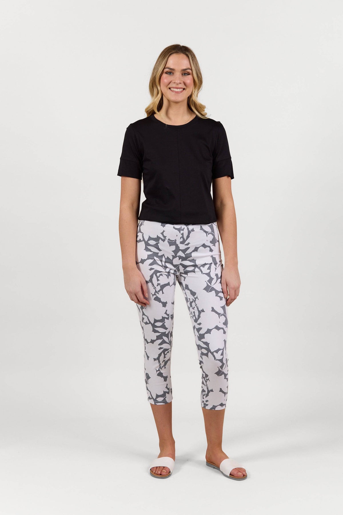 A woman stands smiling in a black short-sleeve top, Vassalli Harbour 7/8 Pant - Floral Gingham, and white slide sandals against a plain white background.