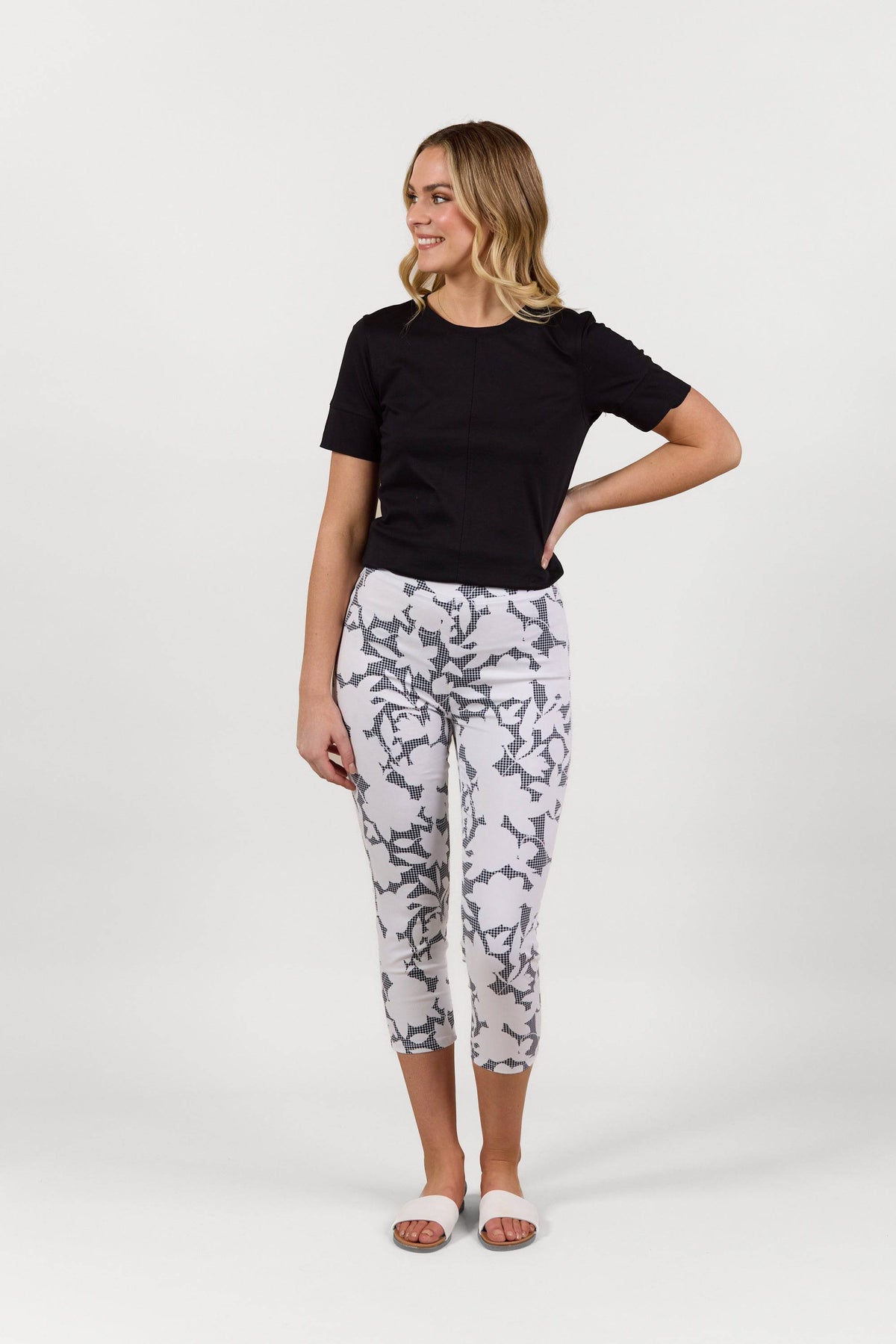 A woman smiles, wearing a black short-sleeve top, Vassalli Harbour 7/8 Pant - Floral Gingham, and white slide sandals, standing against a plain white background.