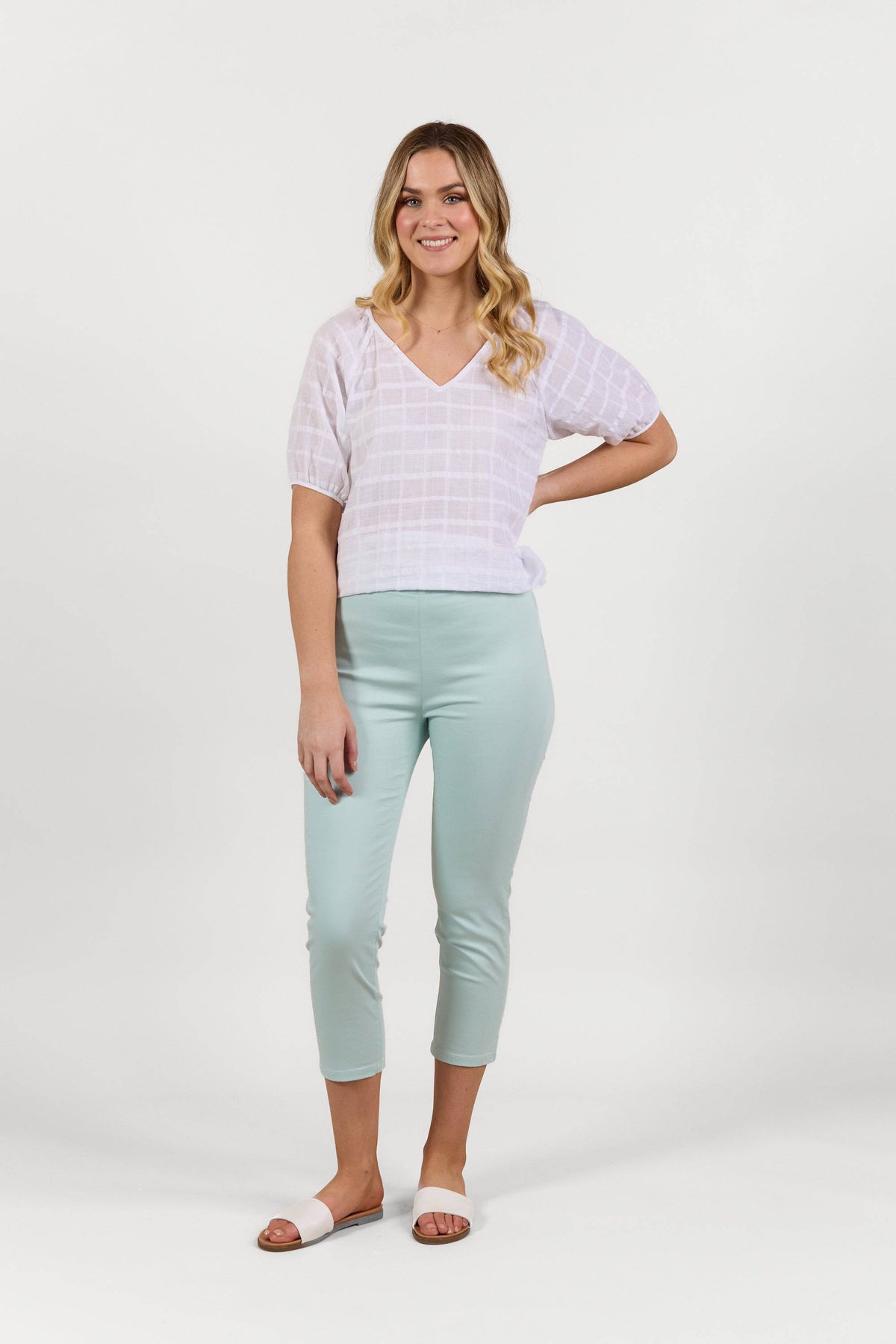 A woman smiles against a white background, wearing a white short-sleeve blouse, Vassalli Harbour 7/8 Pant in Mint, and white slide sandals.