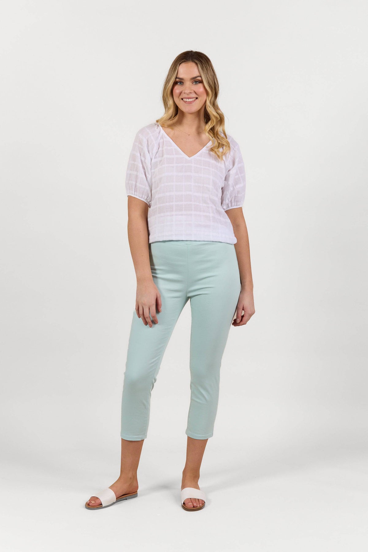 A woman with loose blonde waves smiles against a white background, wearing the Vassalli Harbour 7/8 Pant in Mint, a white short-sleeve V-neck top, and white slide sandals.