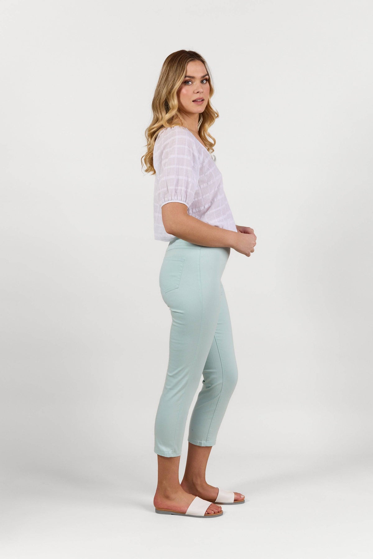 A woman with long blonde hair stands sideways in a white short-sleeve top and Vassalli Harbour 7/8 Pant in Mint, paired with pink slide sandals, against a plain white background.