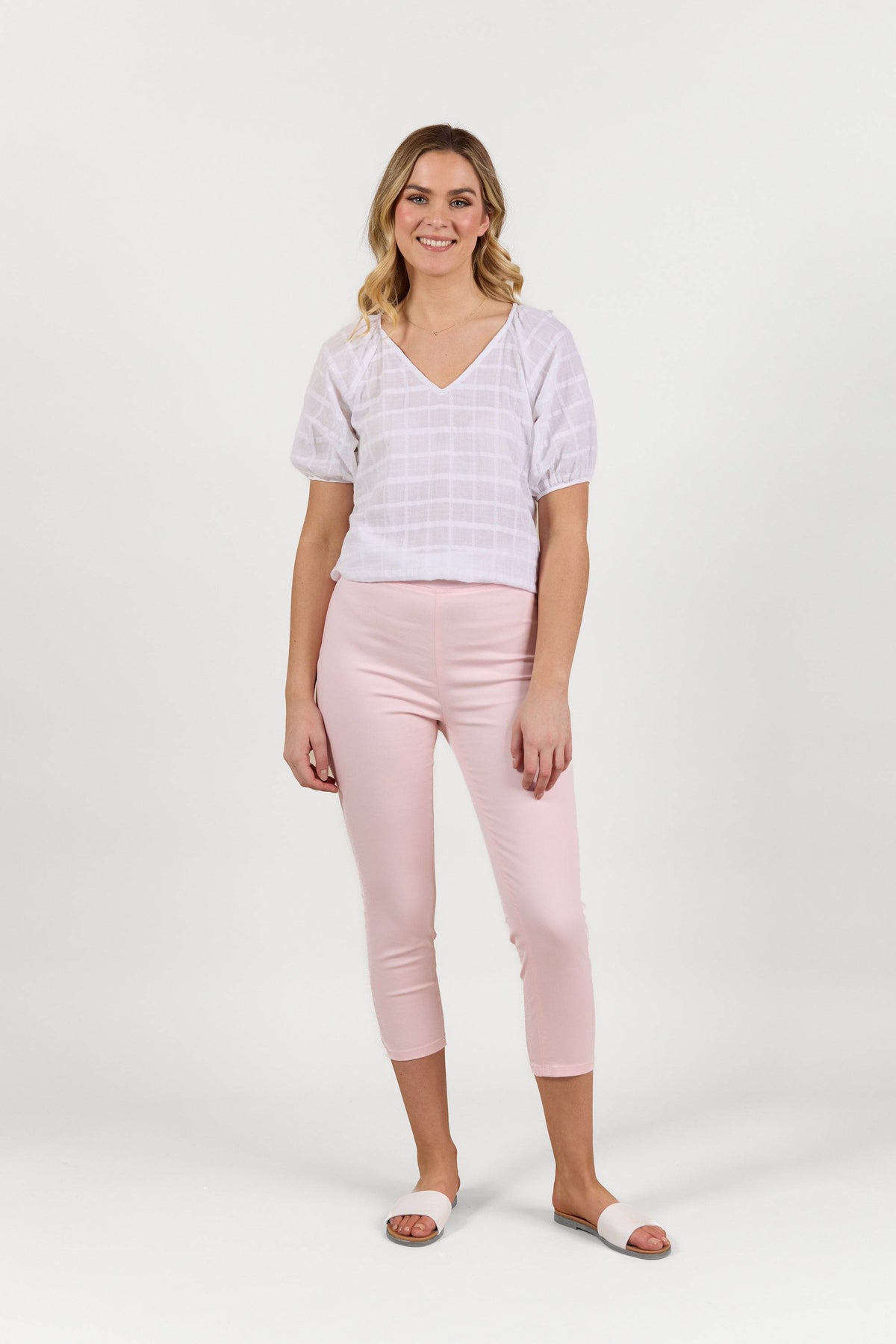 A woman stands smiling against a white background, wearing the Vassalli Harbour 7/8 Pant in Powder Pink, a white short-sleeve blouse, and white slide sandals.