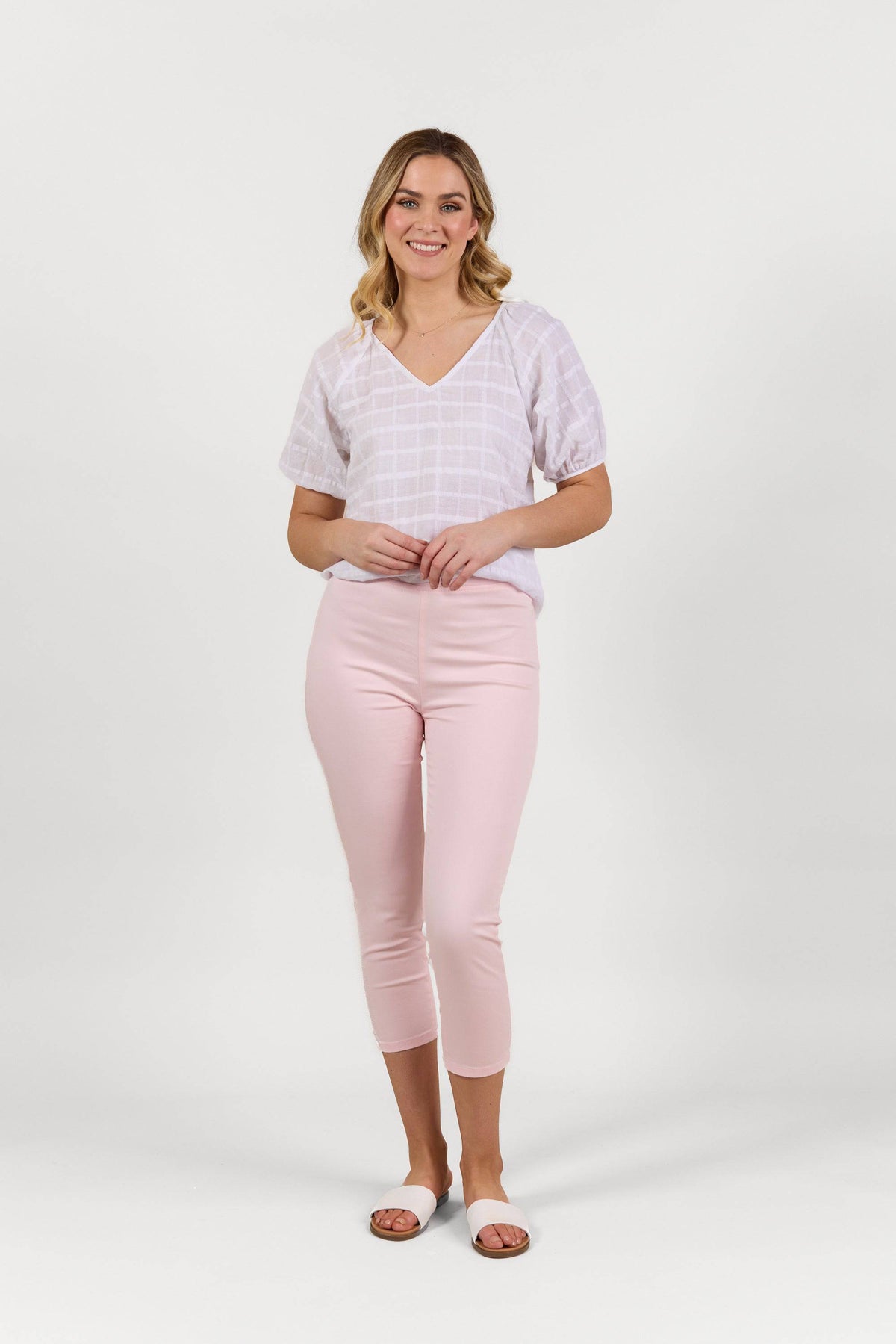 A smiling blonde woman stands against a plain white background wearing Vassalli's Harbour 7/8 Pant in Powder Pink, a white checkered short-sleeve blouse, and white slide sandals.