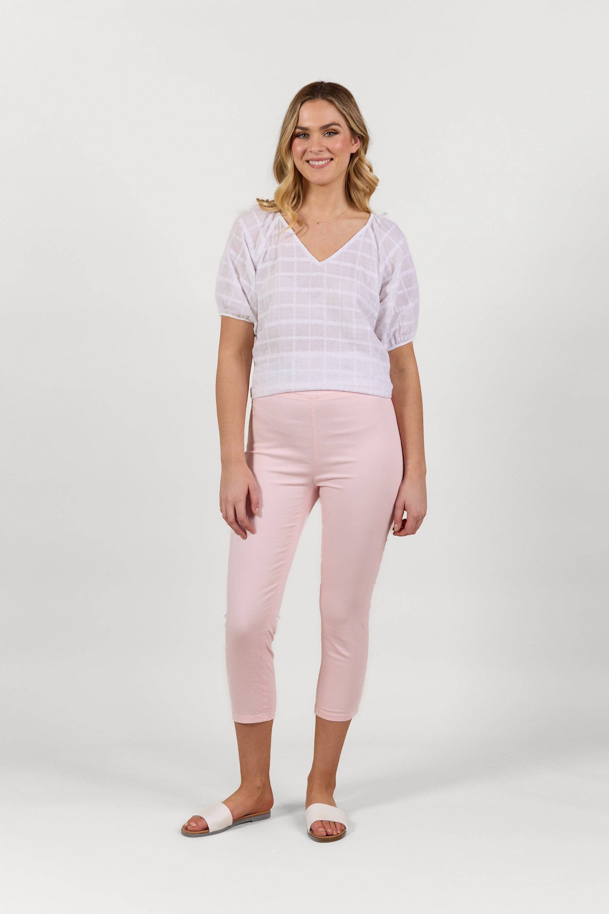 A woman smiles, wearing a white V-neck short-sleeve blouse, Vassalli Harbour 7/8 Pant in Powder Pink, and white slide sandals against a plain white background.