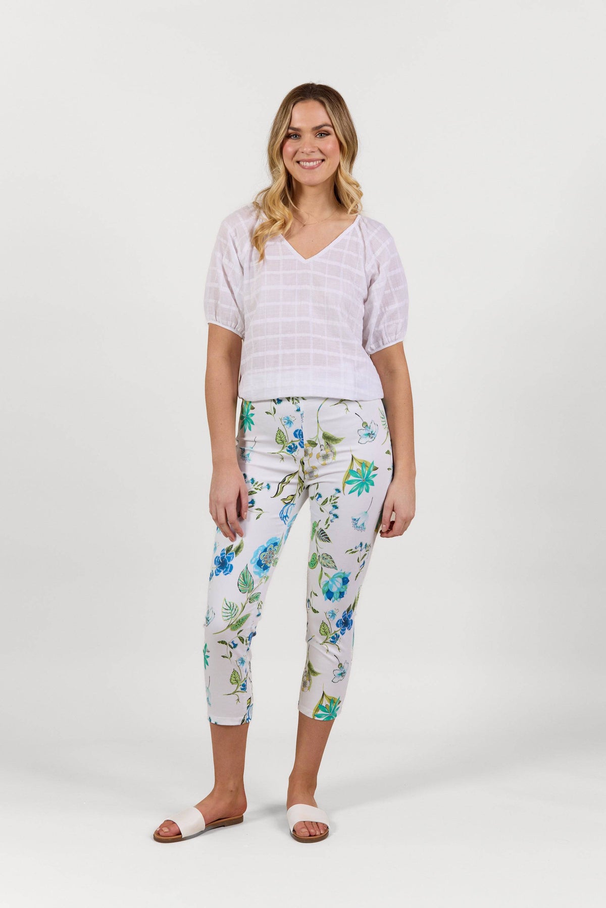 A woman smiles, wearing a white V-neck top, Vassalli Harbour 7/8 Pant in the Santorini print—a blue and green floral design—and white slide sandals. The background is plain white.