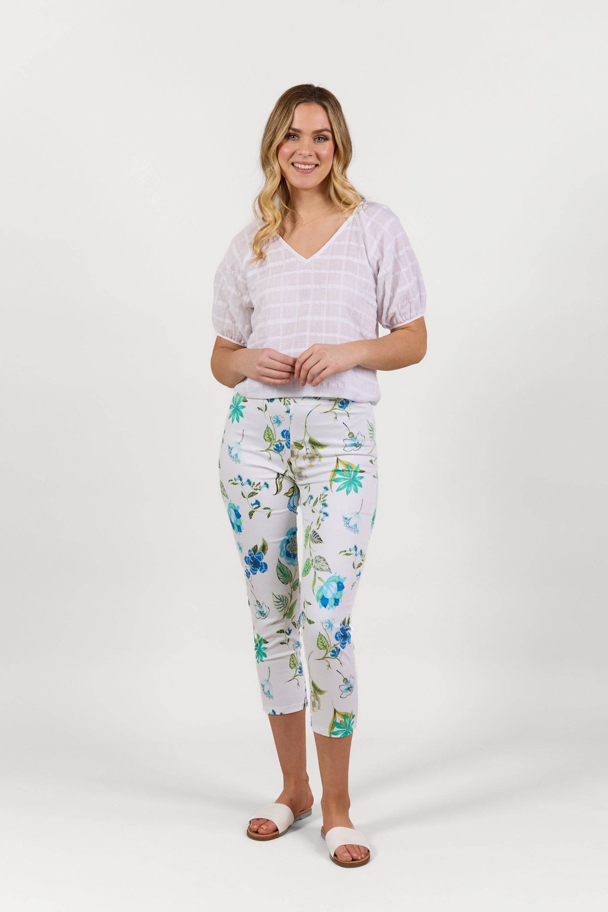 A woman with long blonde hair smiles against a white background, wearing a light purple short-sleeve top, Vassalli Harbour 7/8 Pant - Santorini with a white floral print, and white slide sandals.