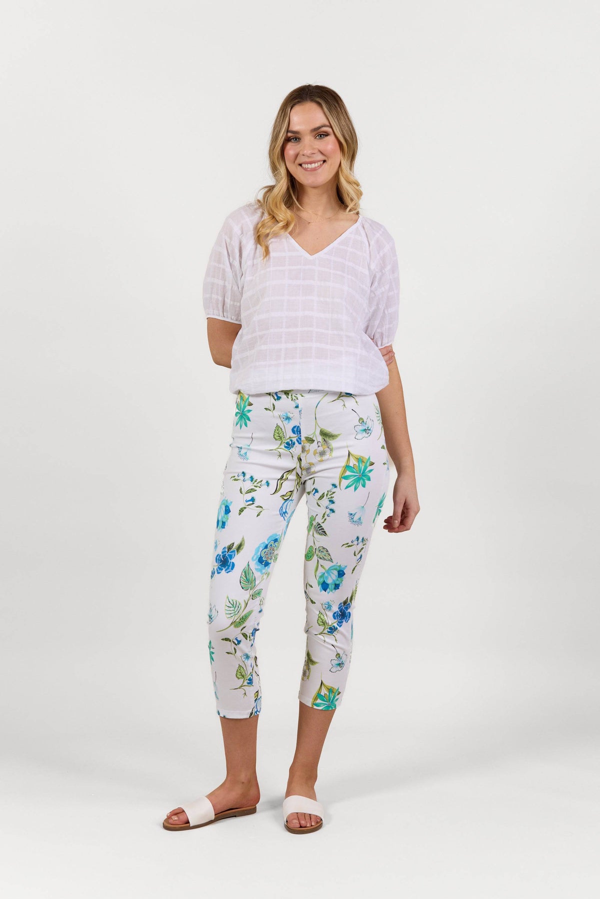 A woman smiles, wearing a white checkered V-neck blouse, Vassalli Harbour 7/8 Pant - Santorini with blue and green floral print, and white slide sandals. The lightweight pull-on pants offer stretchy comfort against a plain white background.