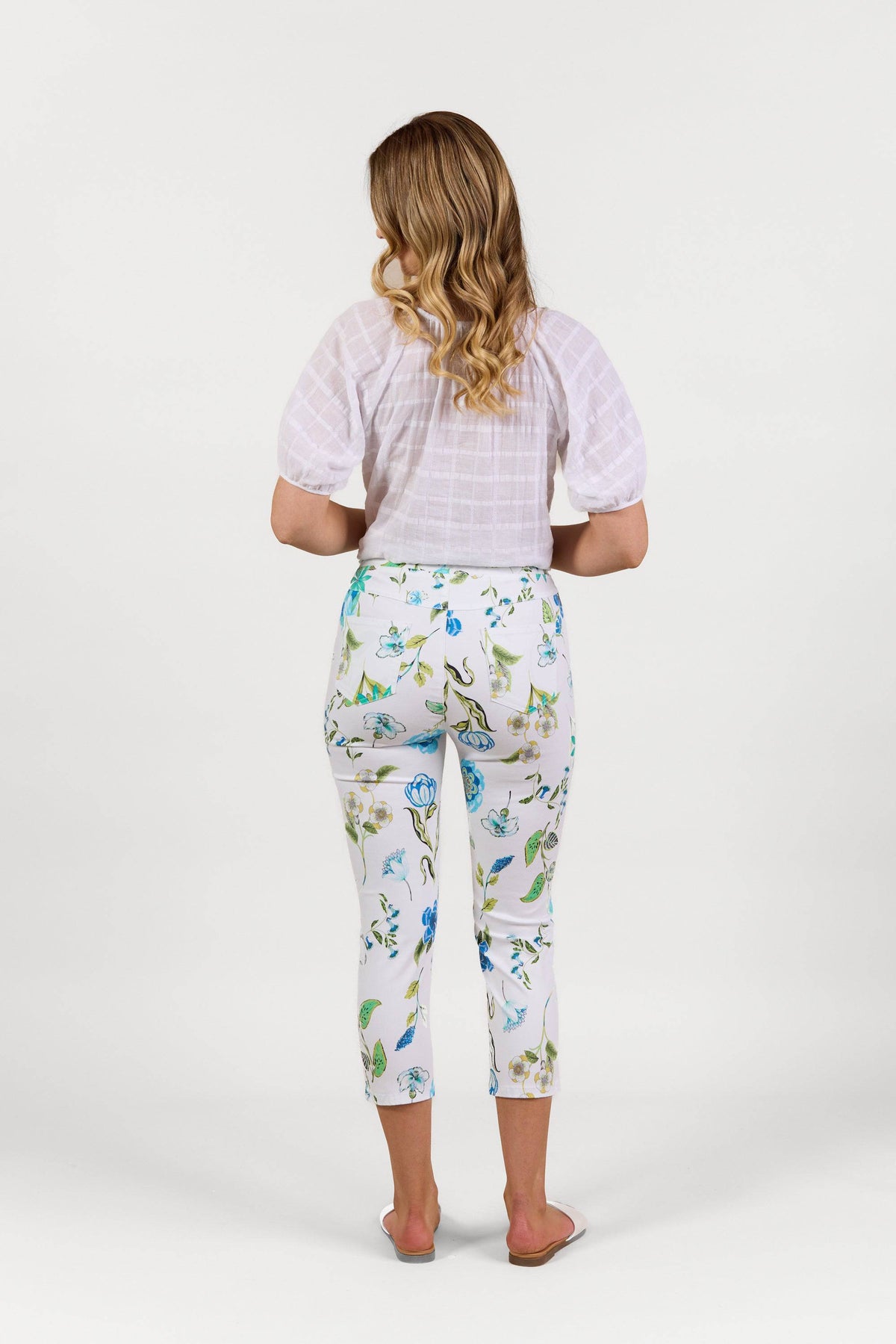 A woman with wavy blonde hair faces away, wearing a white short-sleeve blouse and Vassalli's Harbour 7/8 Pant - Santorini in green and blue. The lightweight pull-on pants pair with white sandals against a plain white background.