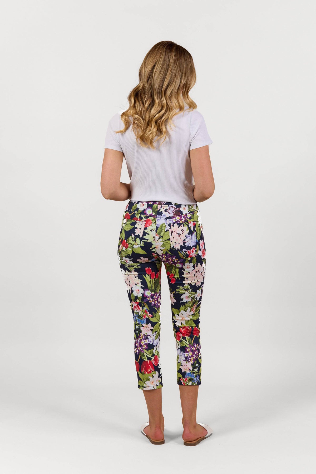 A woman with wavy blonde hair, in a white short-sleeve top and Vassalli Harbour 7/8 Pant - Tulip (printed stretch skinny pants), stands facing away in white sandals against a plain white background.