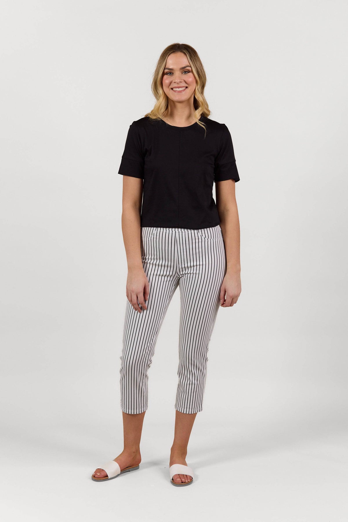 A person smiles against a plain white background, wearing a black short-sleeve top, Vassalli Sierra 7/8 Pant - Black Stripe/White, and white slide sandals.