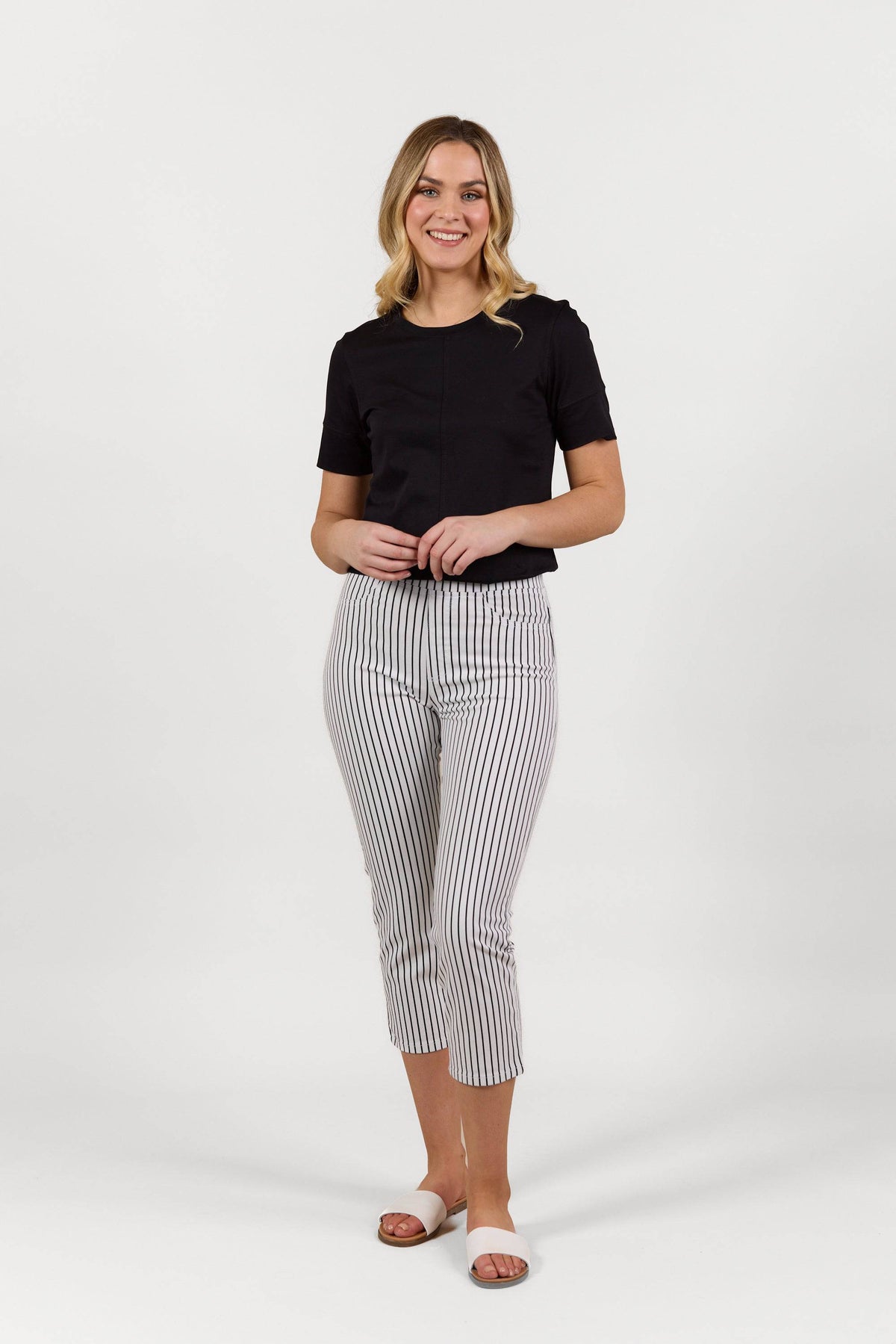 A woman smiles against a white background, wearing a black short-sleeve top, Vassalli Sierra 7/8 Pant in Black Stripe/White, and white slide sandals.
