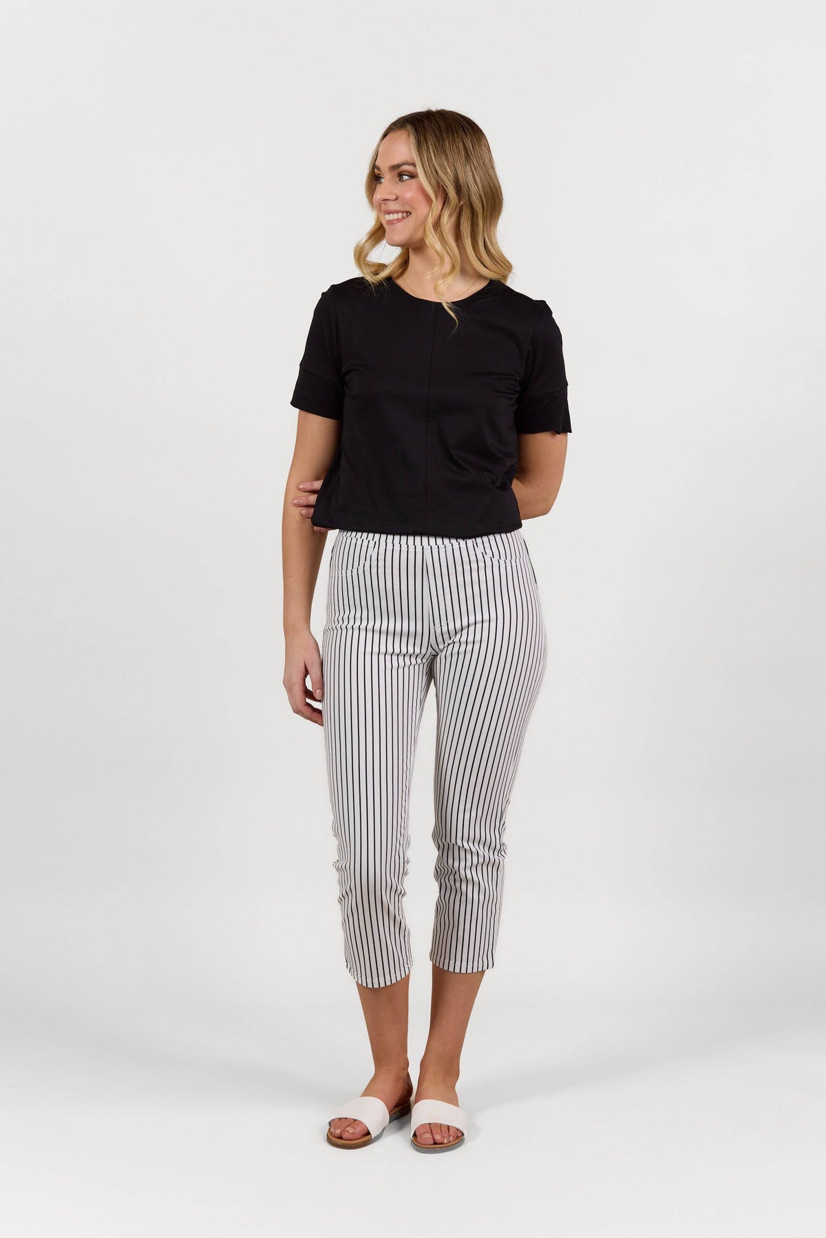 A woman with wavy blonde hair smiles, wearing Vassalli’s Sierra 7/8 Pant in Black Stripe/White, a black short-sleeve top, and white sandals, posing against a plain white background.