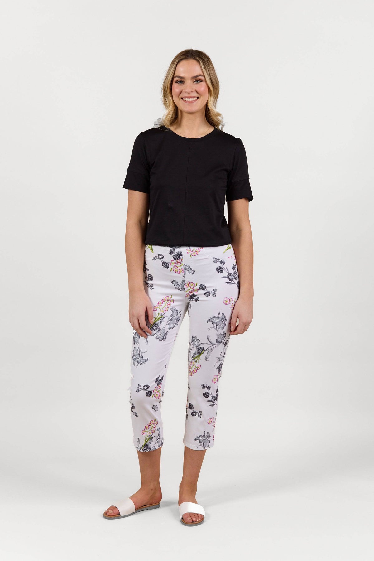 A woman smiles in a short-sleeve black top and Vassalli's Sierra 7/8 Pant - Delphi, featuring a colorful floral pattern in stretch fabric, paired with white slide sandals against a plain white background.
