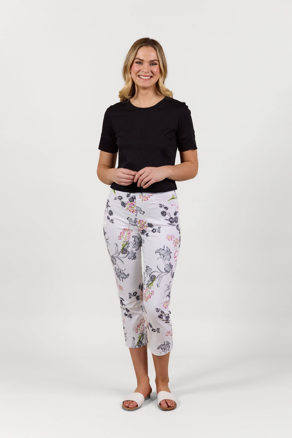 A woman smiles against a plain white background, wearing a black short-sleeve top, Vassalli’s Sierra 7/8 Pant - Delphi with a white floral pattern, and white sandals.