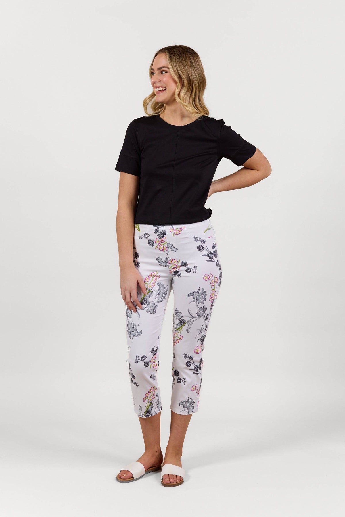 A woman smiles in Vassalli's Sierra 7/8 Pant - Delphi, a colorful floral slim leg stretch pant, paired with a black short-sleeve top and white slide sandals, set against a plain white background.