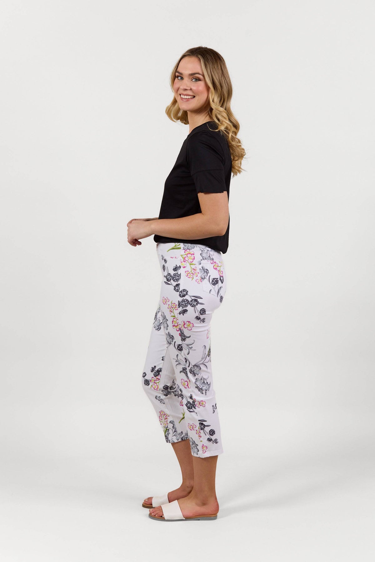 A woman stands sideways, smiling, wearing Vassalli's Sierra 7/8 Pant - Delphi, a white floral slim leg stretch pant, with a black t-shirt and white slide sandals against a plain white background.