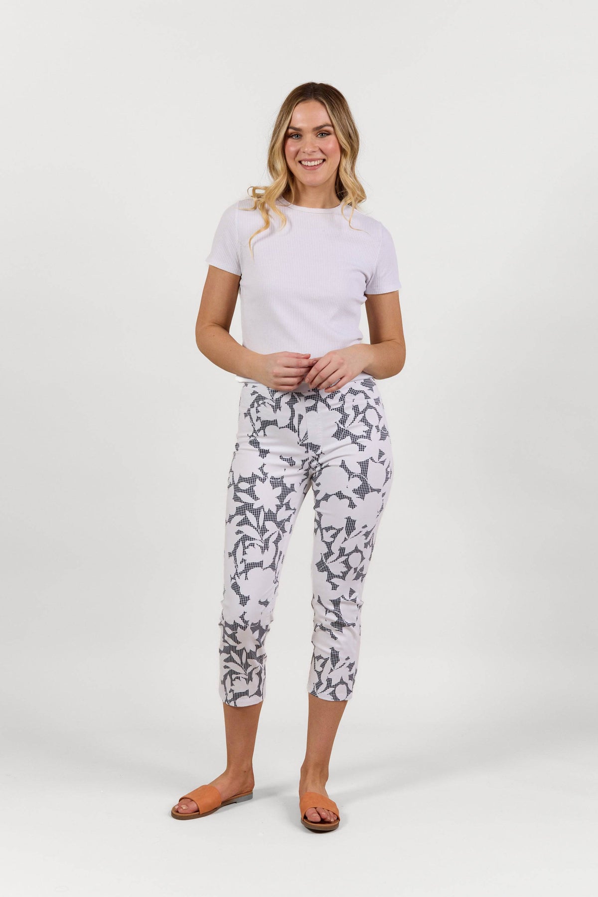 A woman stands smiling against a plain white background, wearing a white short-sleeve top and the Vassalli Sierra 7/8 Pant - Floral Gingham, crafted from stretch fabric, paired with orange sandals.