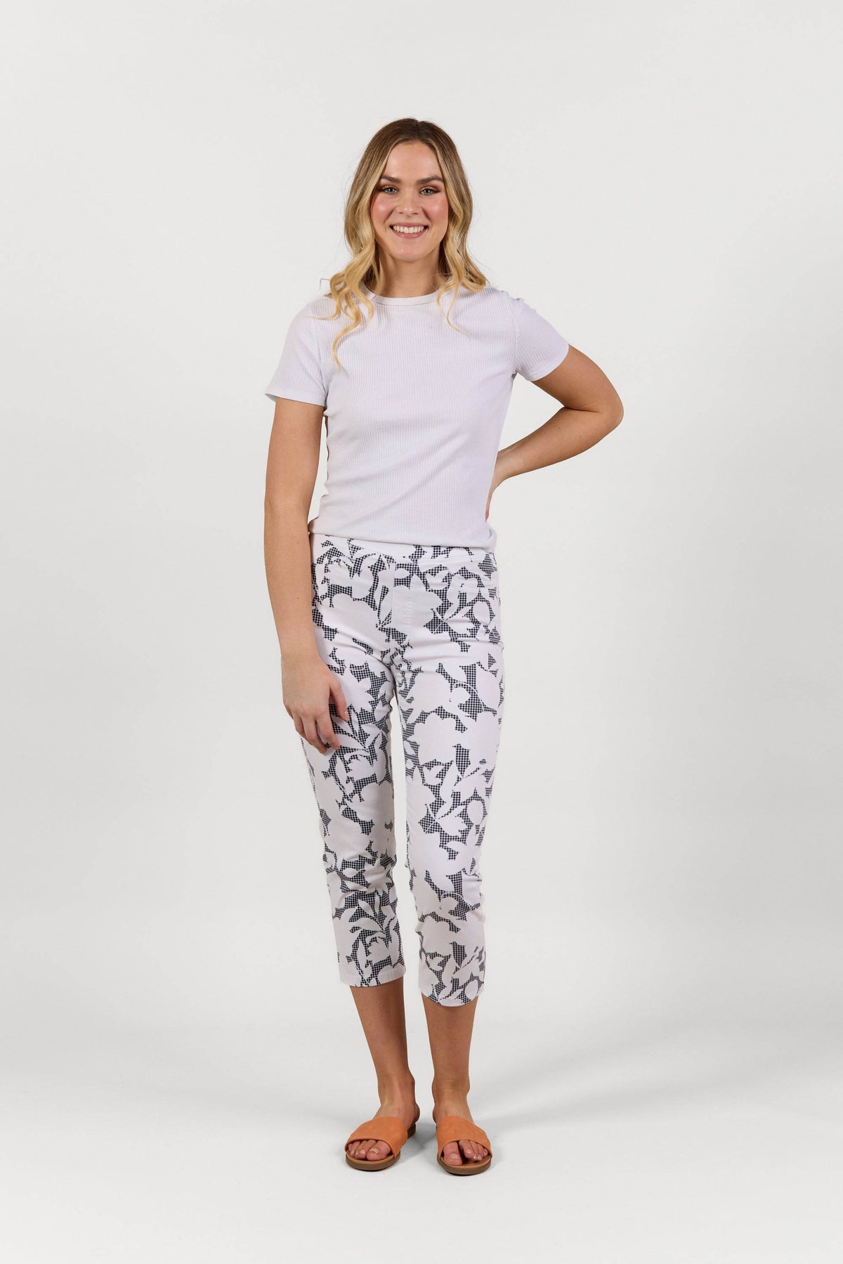 A woman stands smiling against a plain white background, wearing a white short-sleeve shirt, brown sandals, and the Vassalli Sierra 7/8 Pant in Floral Gingham print, crafted from stretch fabric.
