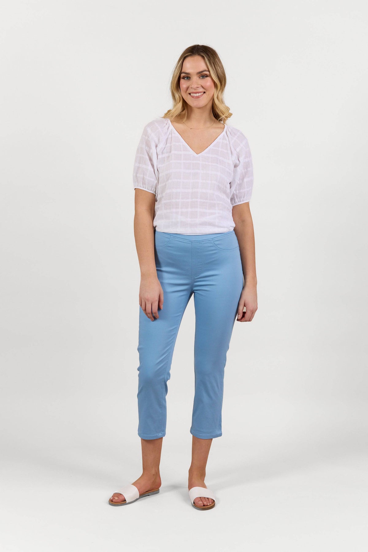 A woman stands against a plain white background, smiling. She is wearing a white short-sleeve blouse, Vassalli’s Sierra 7/8 Pant in Fresh Blue, and white slide sandals.