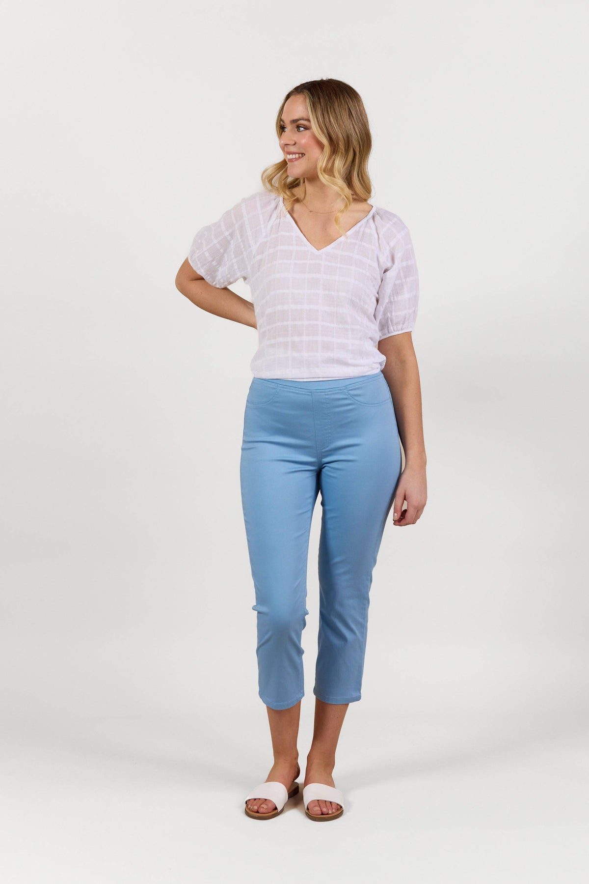 A woman smiles, wearing a sheer white short-sleeve top, Vassalli Sierra 7/8 Pant in Fresh Blue, and white slide sandals, against a plain white background.