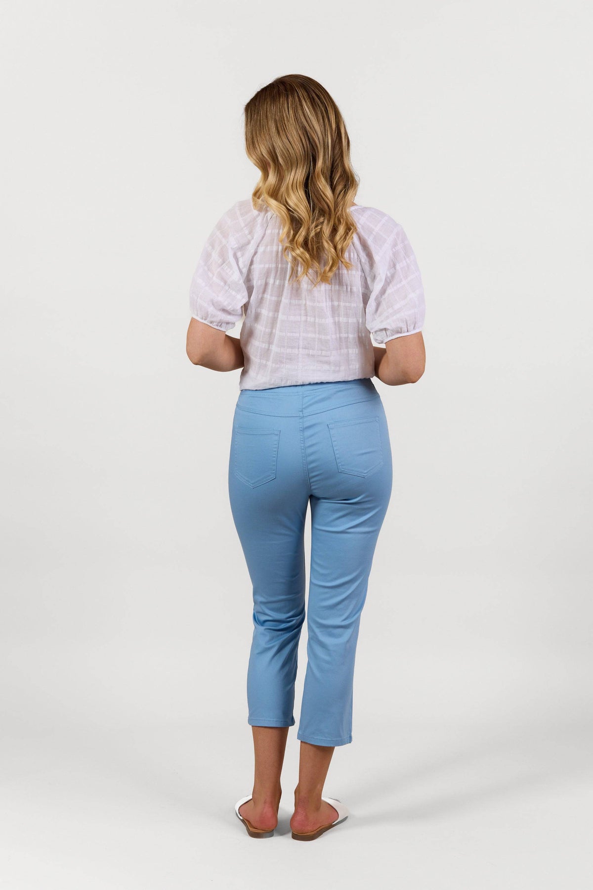 A woman with wavy blonde hair stands facing away, wearing a white short-sleeve blouse and Vassalli Sierra 7/8 Pant in Fresh Blue, styled with white flat sandals against a plain white background.