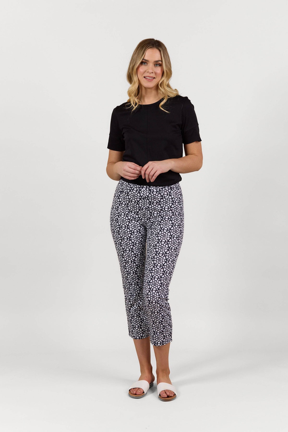 A woman smiles against a white background, wearing Vassalli's Sierra 7/8 Pant - Hearts with a black short-sleeve top and white slide sandals. Her lightweight pull-on pants feature a black-and-white heart pattern.