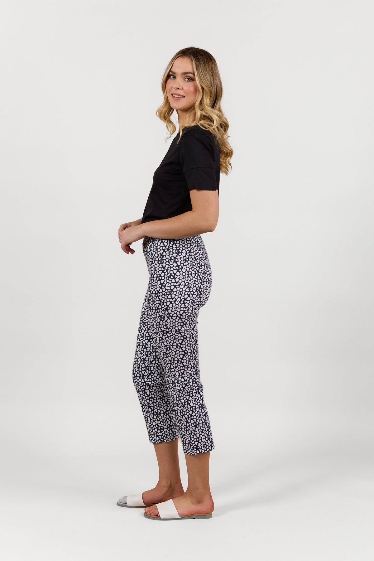 A woman stands sideways against a white background, wearing a black t-shirt and Vassalli Sierra 7/8 Pant - Hearts. She completes her look with slide sandals, her wavy blonde hair framing a gentle smile.