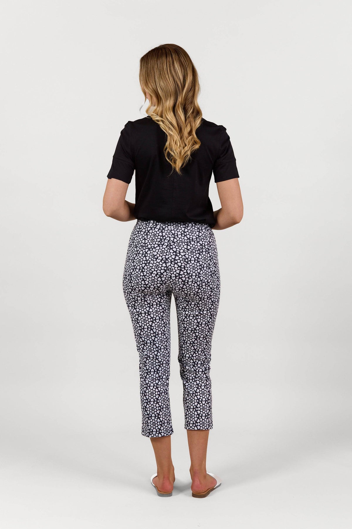 A woman with wavy blonde hair, in a black short-sleeve top and Vassalli Sierra 7/8 Pant - Hearts, stands barefoot facing away against a plain white background. These lightweight pull-on pants enhance her stylish look.