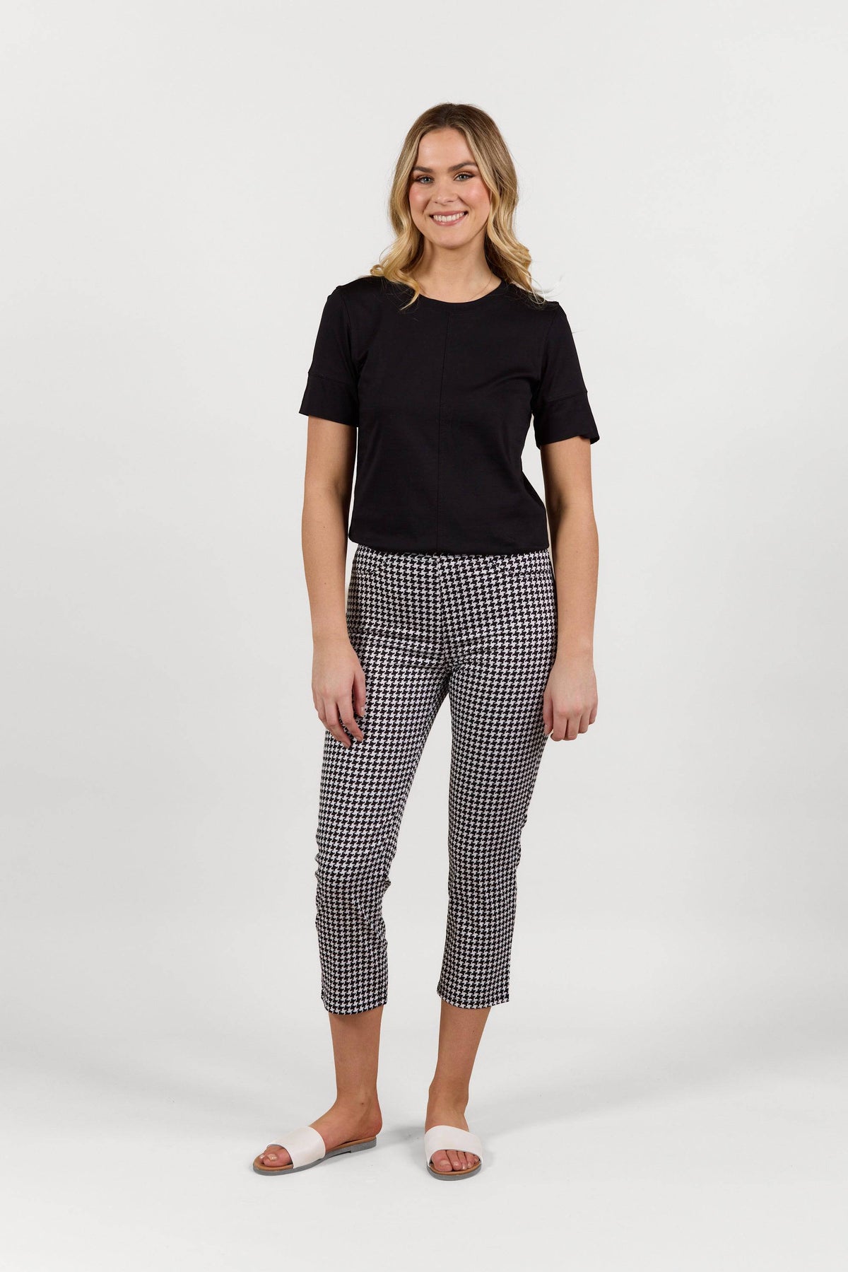A woman smiles against a plain white background, wearing a black short-sleeve top, Vassalli Sierra 7/8 Pant in houndstooth, and white slide sandals.