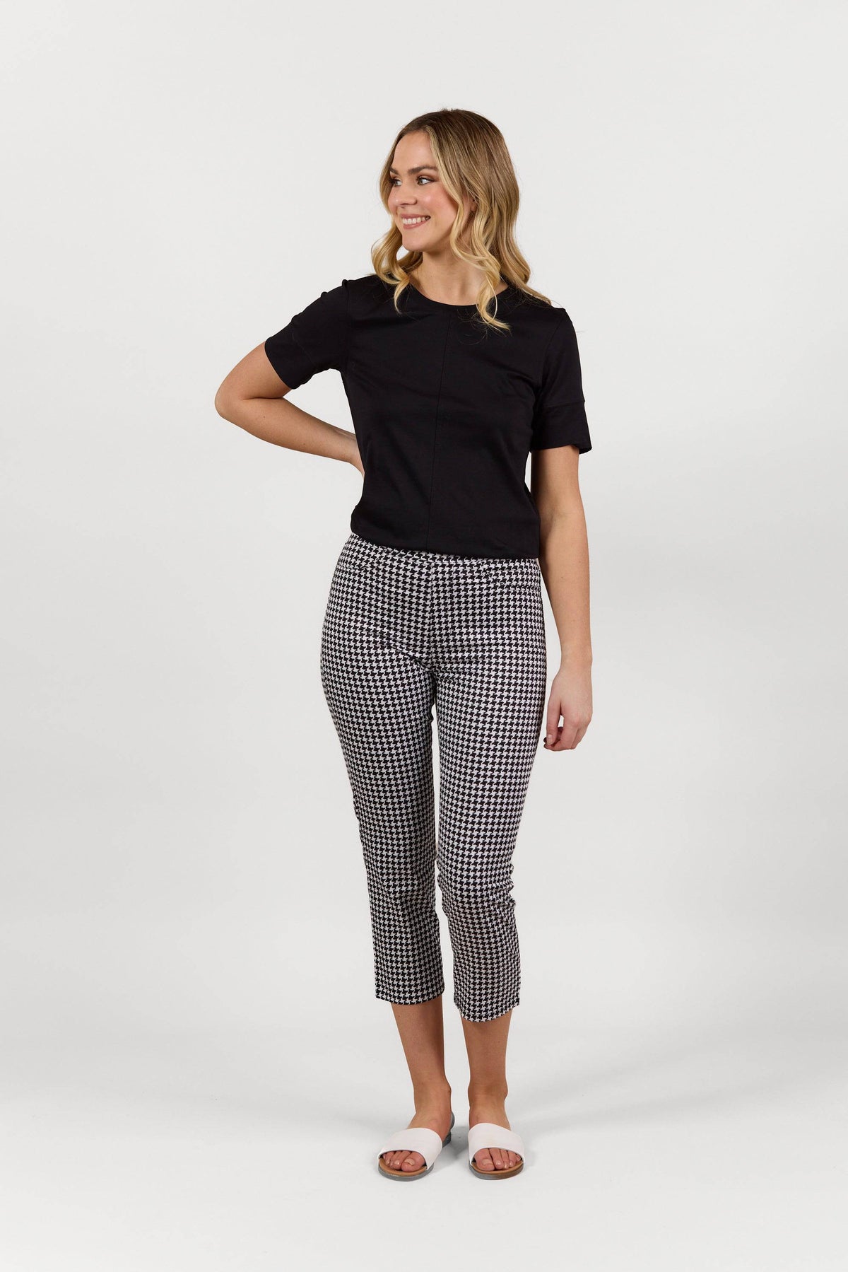 A person stands against a white background, wearing a black short-sleeve top and Vassalli Sierra 7/8 Pant - Houndstooth with white sandals. They are smiling, looking to the side, with one hand resting by their side.