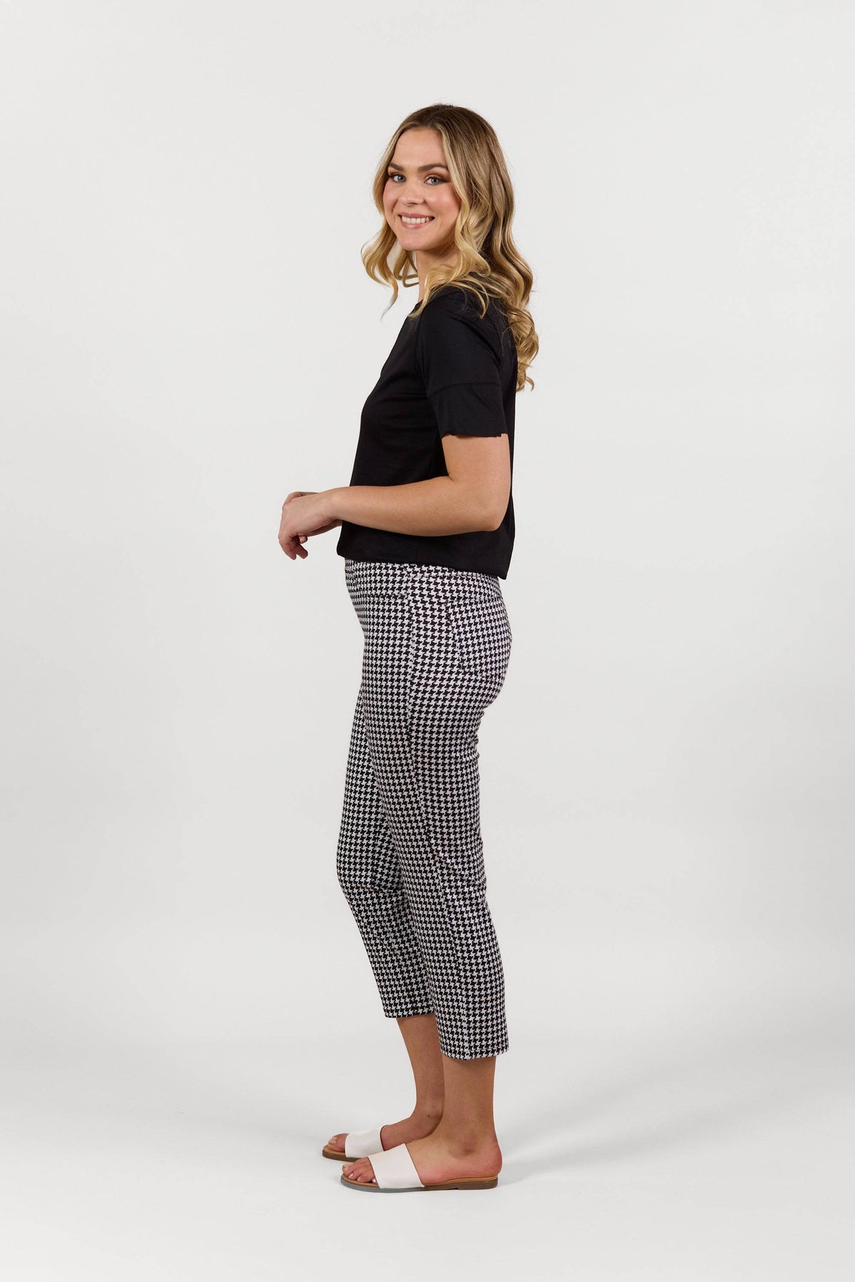 A woman stands sideways, smiling, in a black short-sleeve top and Vassalli Sierra 7/8 Pant - Houndstooth with white slide sandals against a plain white background.