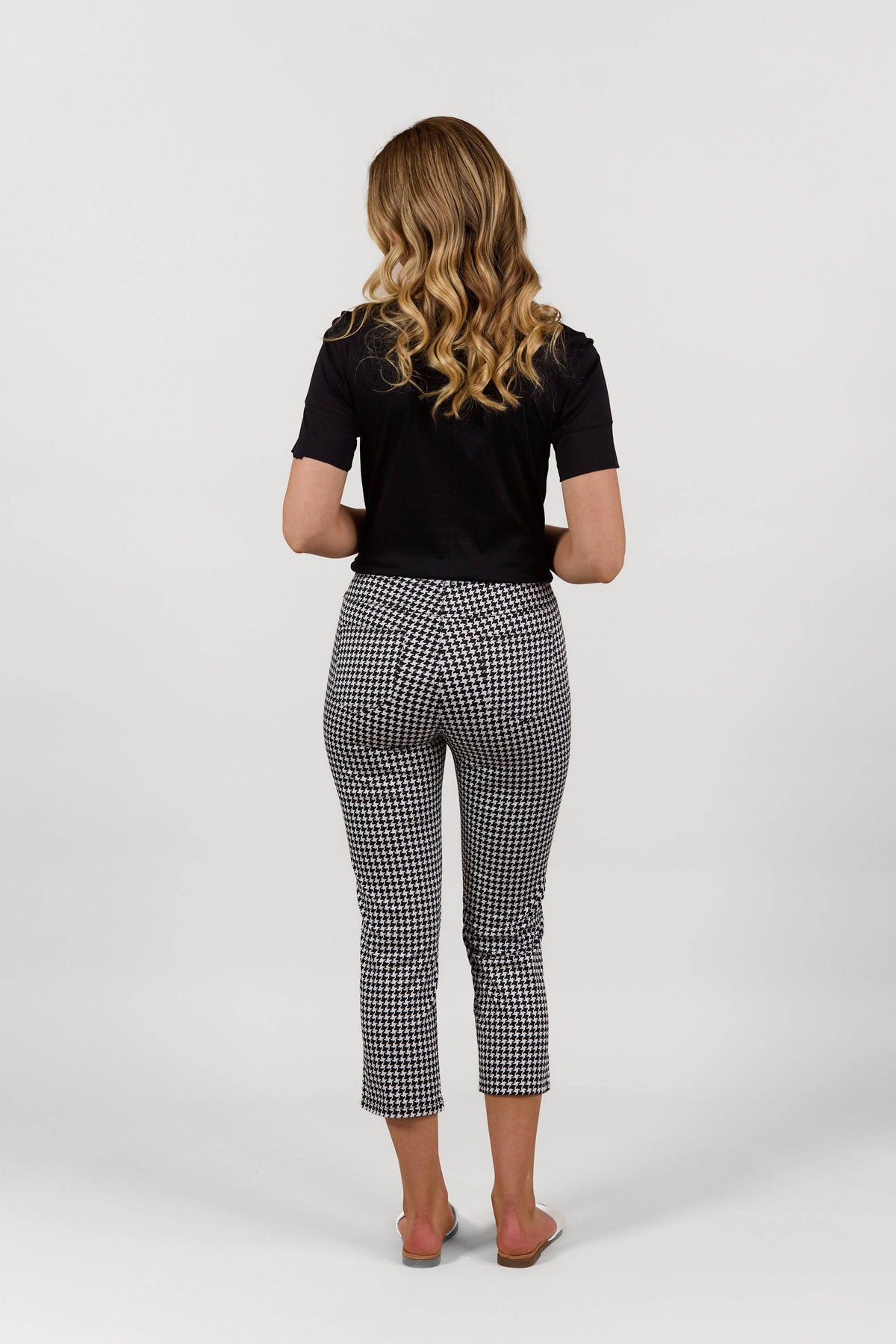 A woman with long wavy blonde hair stands facing away, wearing a black short-sleeve top and Vassalli Sierra 7/8 Pant - Houndstooth, paired with light-colored slides, against a plain white background.