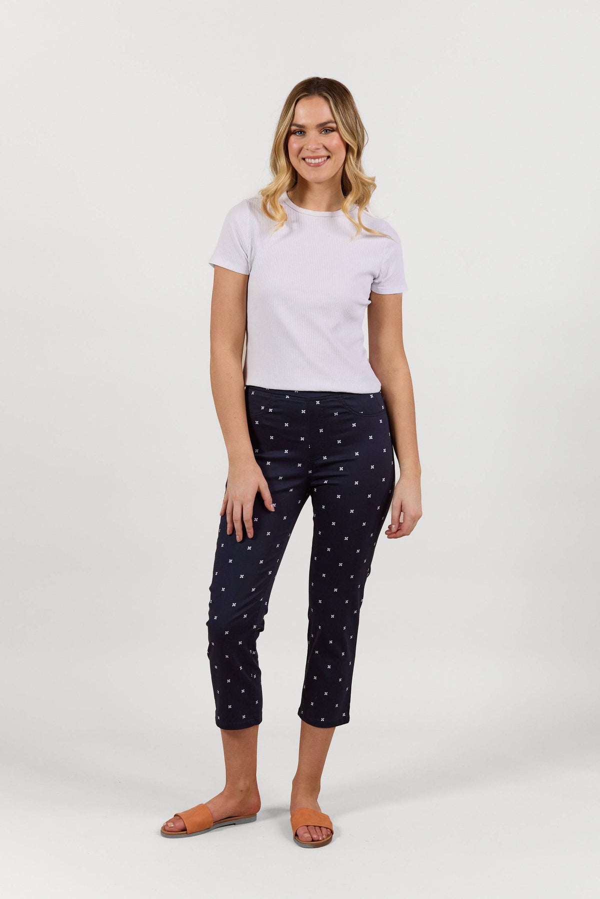 A woman smiles against a white background, wearing a short-sleeve white tee, Vassalli Sierra 7/8 Pant - Navy Daisy with a small white print in stretch fabric, and tan slide sandals.
