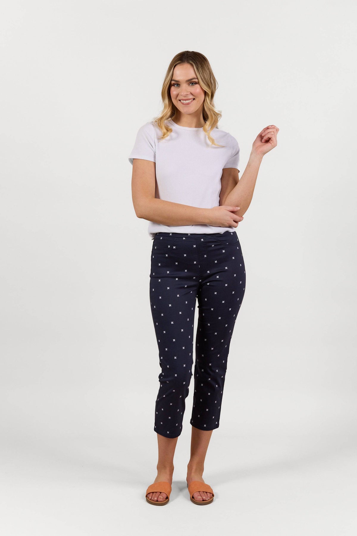 A woman smiles against a white background, wearing a short-sleeve white top, Vassalli Sierra 7/8 Pant - Navy Daisy with a white daisy print, and orange sandals.