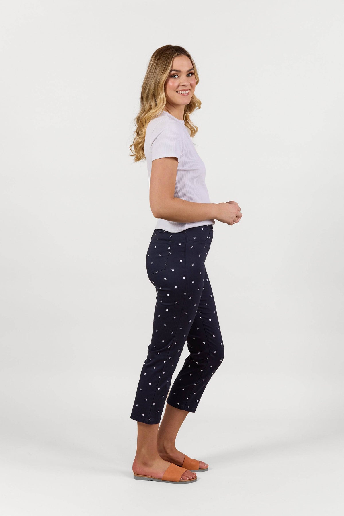 A woman with long blonde hair, wearing a white short-sleeve top and Vassalli's Sierra 7/8 Pant - Navy Daisy, stands sideways in orange sandals and smiles against a plain white background.