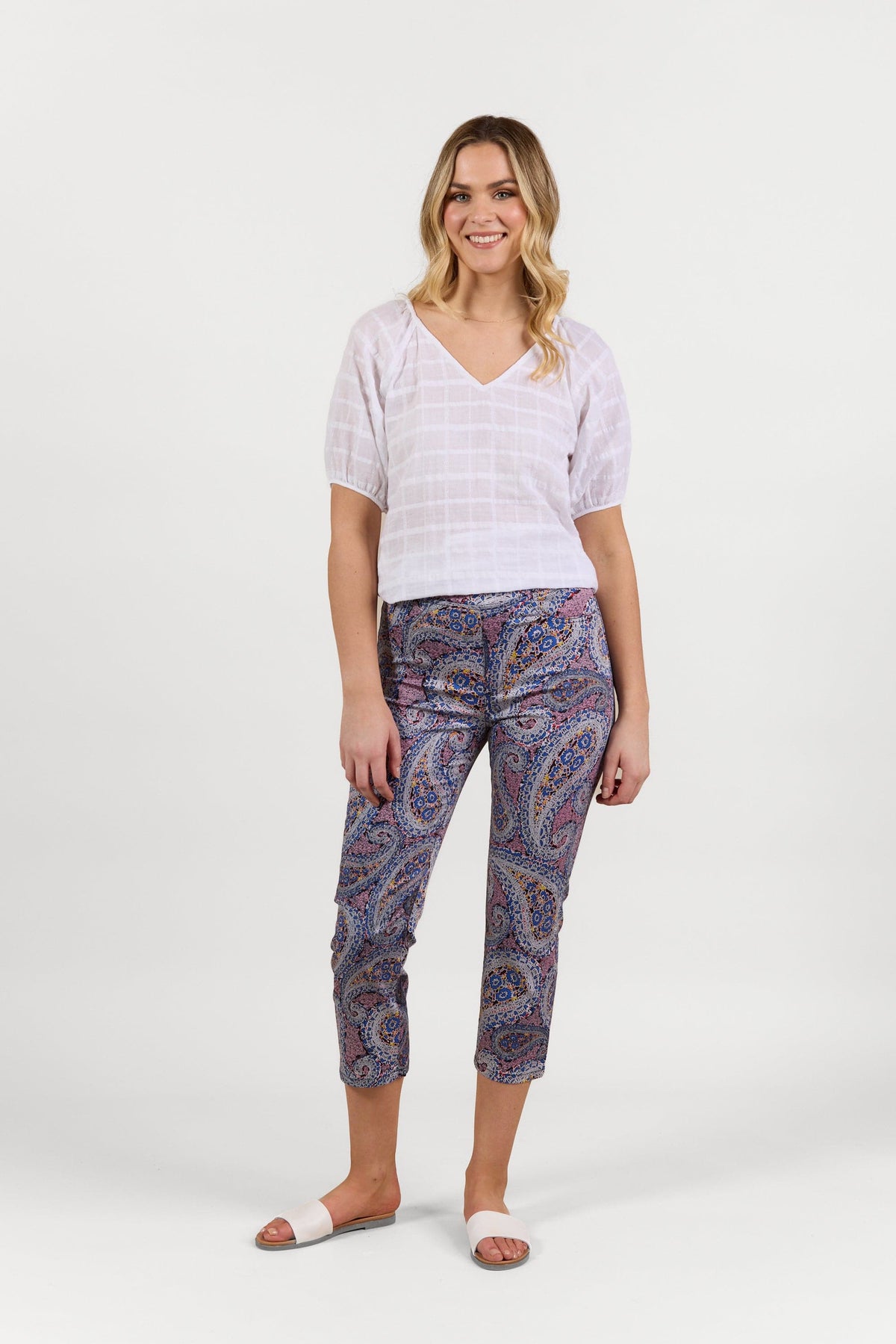 A woman smiles, wearing a white short-sleeve blouse, Vassalli Sierra 7/8 Pant - Paisley, and white slide sandals against a plain white background.